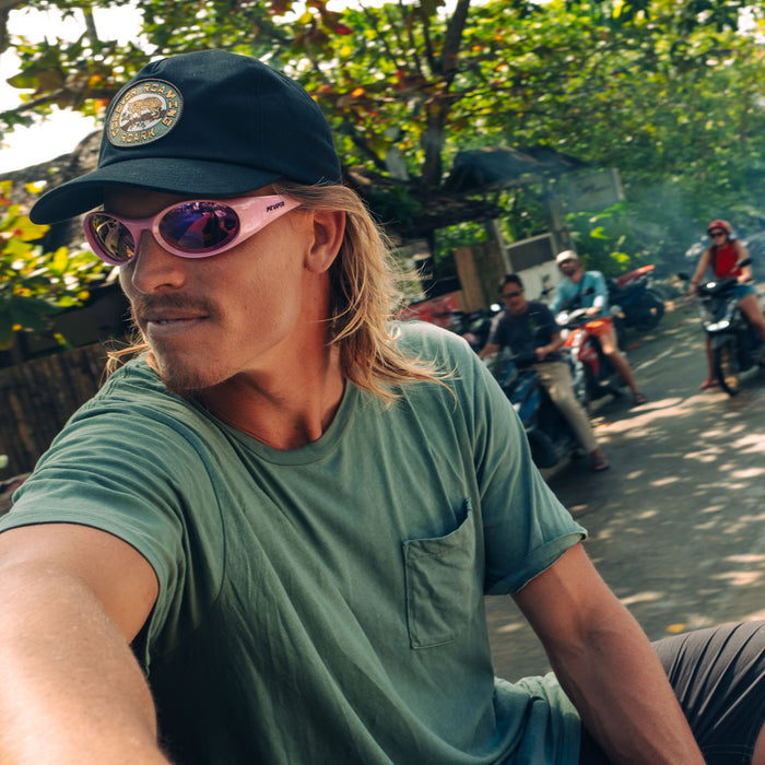 Man wearing washed slate green short sleeve tee with chest pocket, styled with black cap and pink sunglasses outdoors