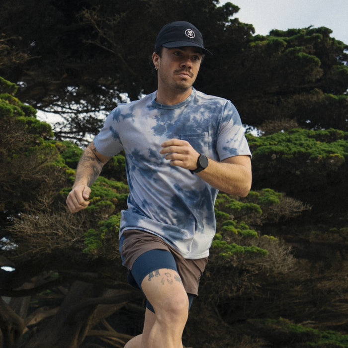 Man showcasing active lifestyle by wearing tie-dye pocket tee paired with shorts, accessorized with a hat, outdoors near the coast