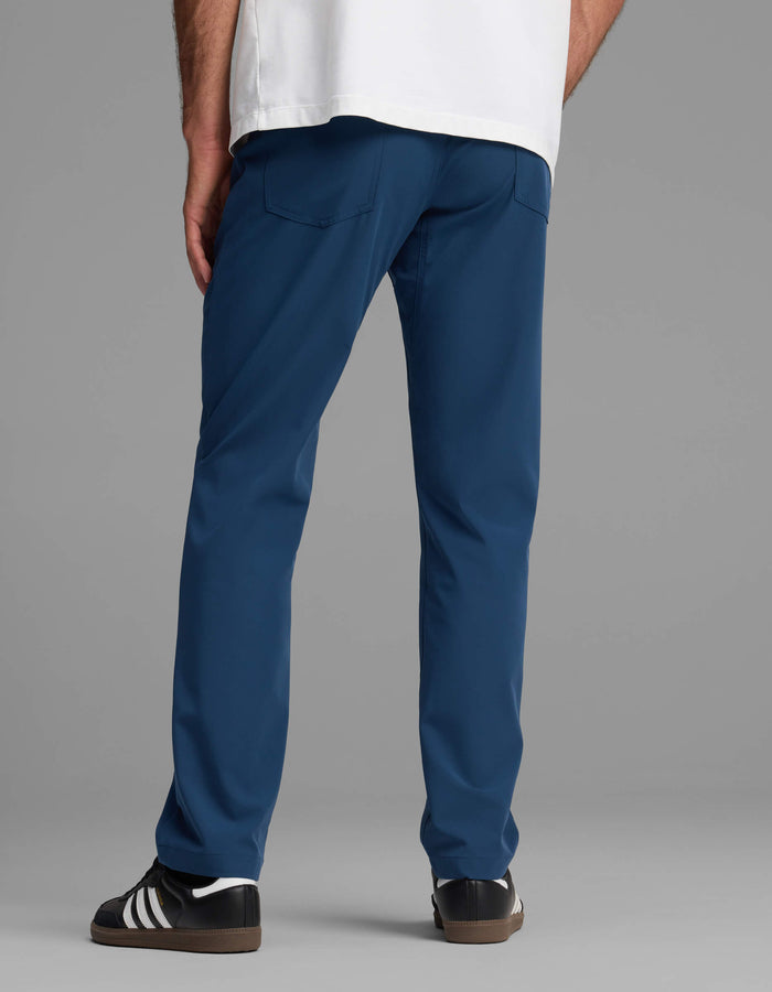 A person stands facing away against a plain gray background, wearing Cozy Earth's Men's Everywhere Pant - Relaxed Fit in blue, paired with a white shirt and black sneakers with white stripes.|Color:Obsidian