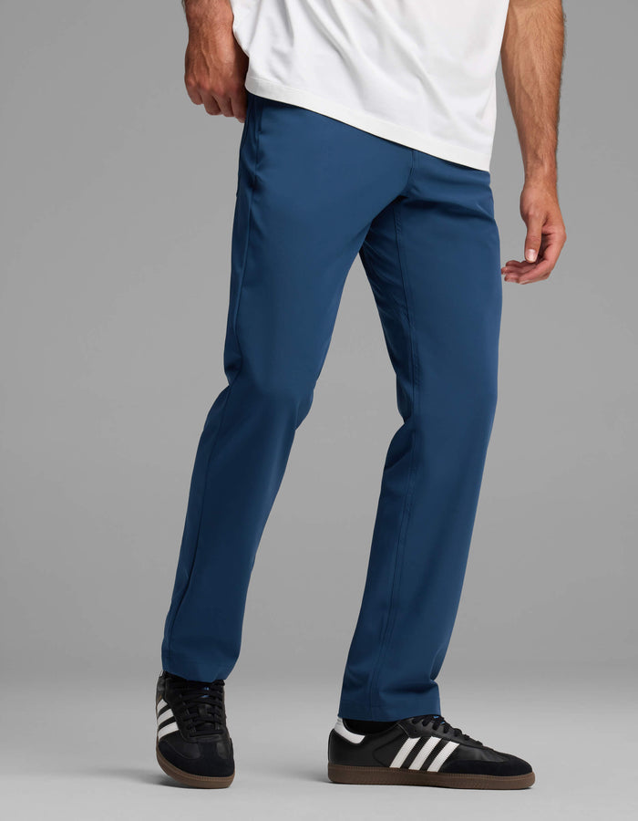 A person models Cozy Earth's Men's Everywhere Pant - Relaxed Fit in blue with a white shirt and black Adidas sneakers, standing against a gray background. The image is cropped at the shoulders.|Color:Obsidian