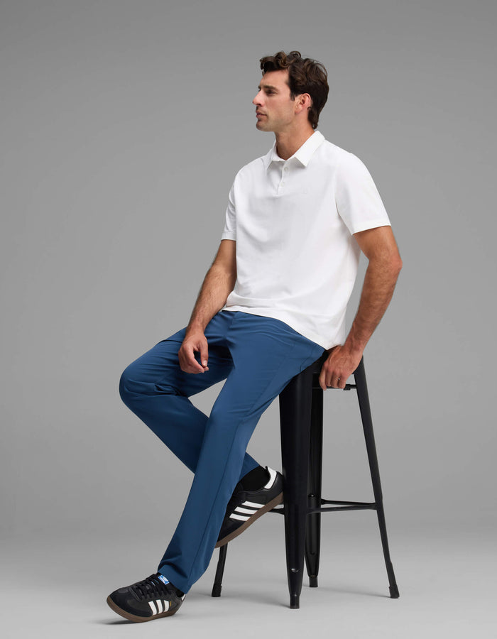 A man sits on a tall black stool against a plain gray background, wearing Cozy Earth's Men's Everywhere Pant - Relaxed Fit with a white polo shirt and black Adidas sneakers, looking to the side with a relaxed expression.|Color:Obsidian