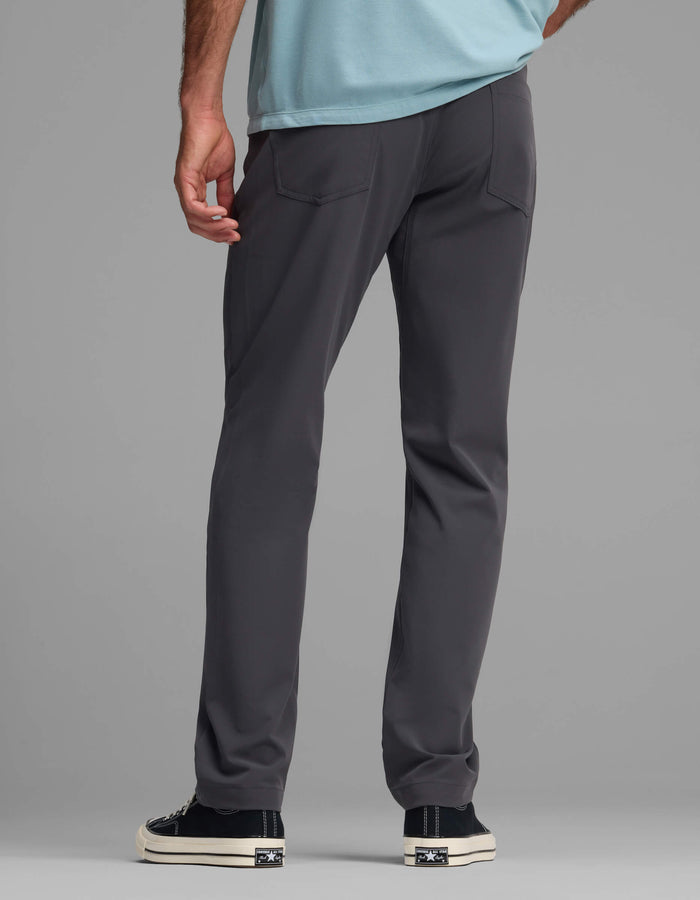 A person is seen from behind wearing the Cozy Earth Men's Everywhere Pant - Relaxed Fit in dark gray, paired with a light blue shirt and black-and-white sneakers, standing against a plain gray background.|Color:Graphite