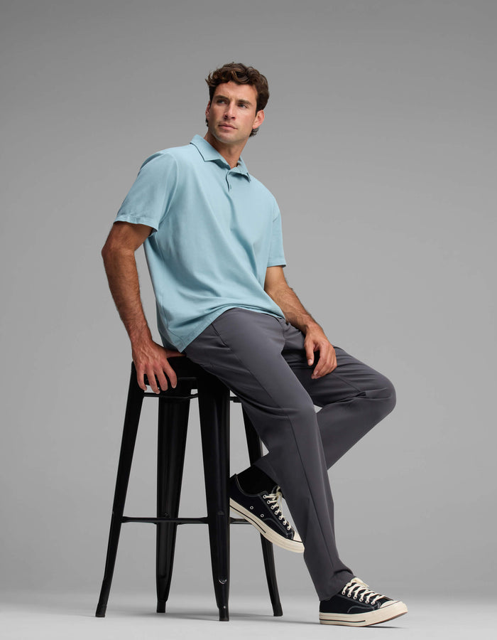 A man in Cozy Earth's Men's Everywhere Pant - Relaxed Fit, paired with a light blue polo and black sneakers, sits on a tall black stool against a plain gray background, gazing off to the side.|Color:Graphite