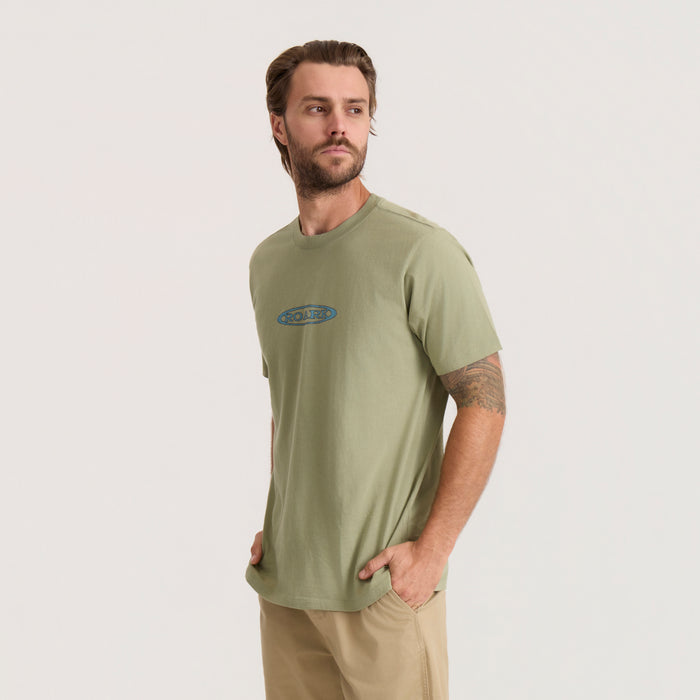 Dusty sage short sleeve t-shirt with blue oval logo on chest, worn by male model with hands in pockets
