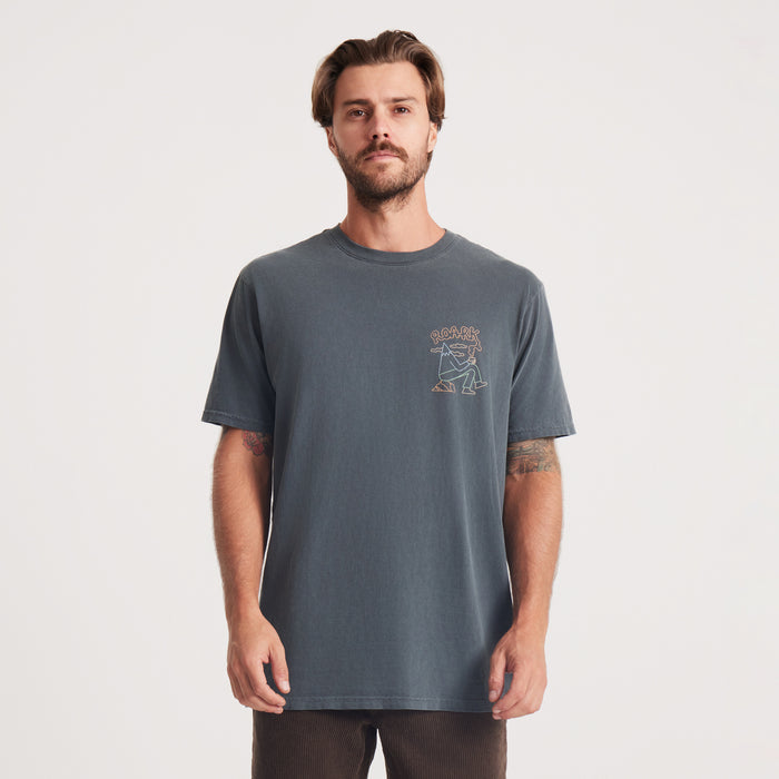 A man wears a cream Roark t-shirt, standing in a relaxed pose, showcasing a minimalist design that’s perfect for layering or solo wear.