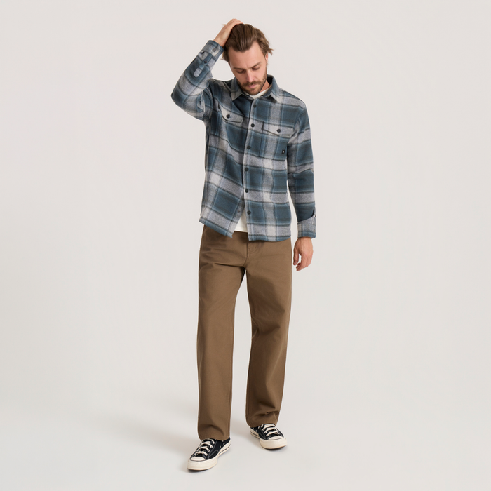 Man wearing dark slate plaid flannel shirt with brown pants and black sneakers, hand to head in casual pose