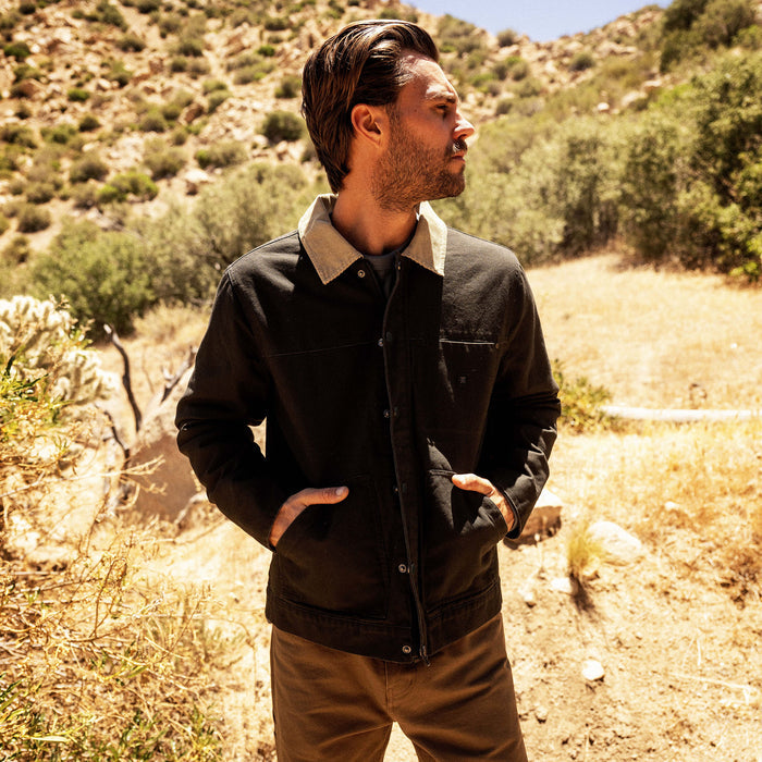 Man wearing black waxed canvas jacket with tan corduroy collar and chest pocket in desert landscape setting