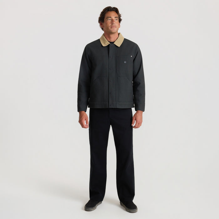 Man wearing black waxed canvas jacket with tan collar and front zip, paired with black pants, standing against white background