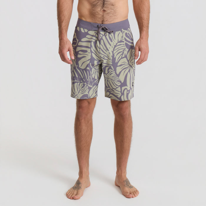 Surfer preparing to hit the waves, sporting durable Roark boardshorts and carrying a sleek surfboard under a bright sky.