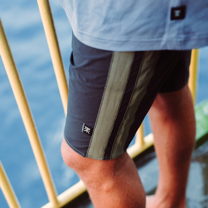 Passage Panel Boardshorts in black with olive stripe detail, worn on model near water with oars visible