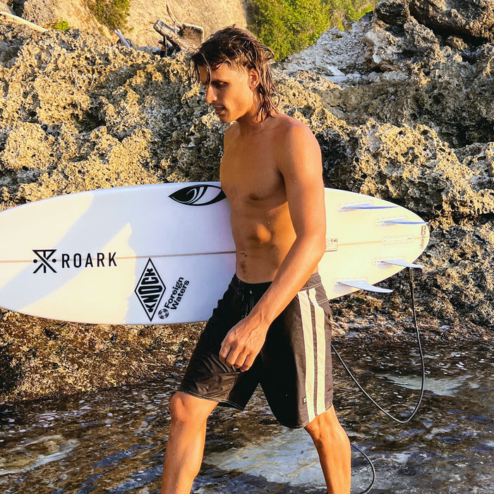 Male surfer wearing black and white Roark Passage Panel boardshorts, holding white surfboard at rocky coastal beach
