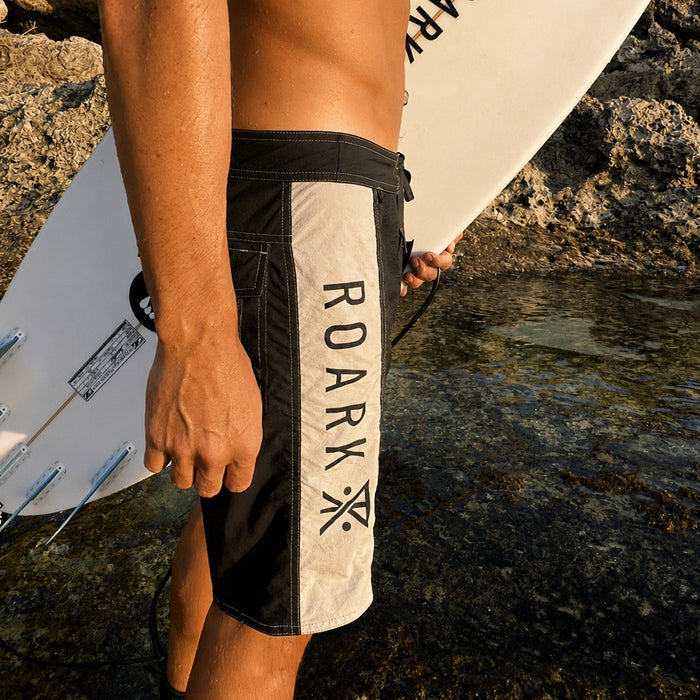 Roark Passage Panel boardshorts in black and white worn by model at rocky beach, showing cream panel with brand text