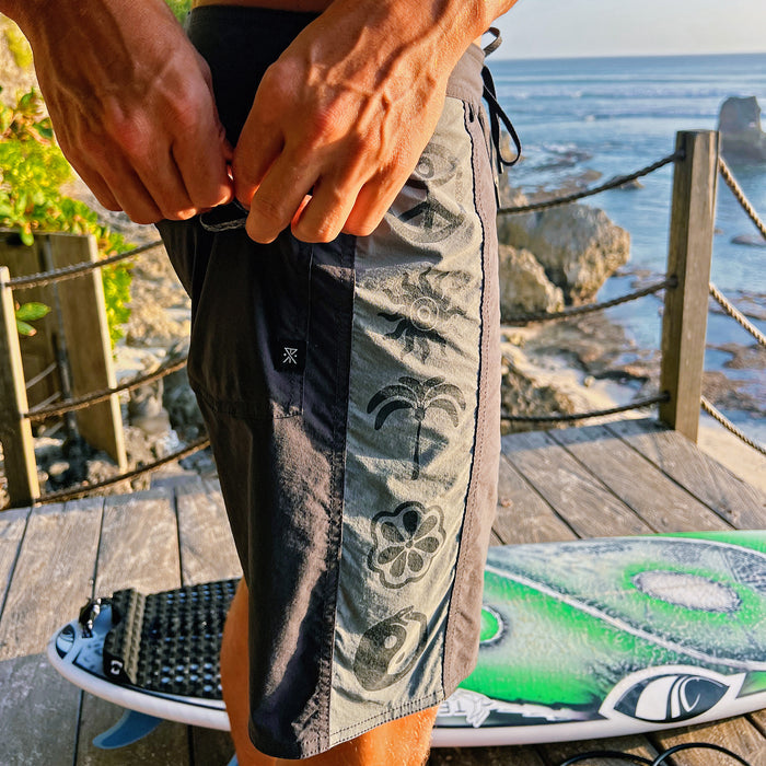 Passage Panel Boardshorts in ebony with tropical print, worn on model at seaside deck overlooking ocean rocks and water