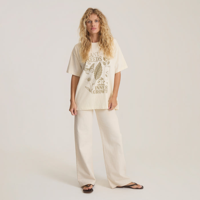 Cream oversized tee with botanical plant seeds graphic and tan text worn by model with matching cream pants