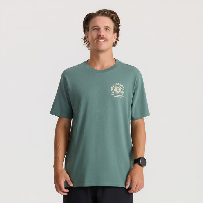 Sage green short sleeve tee with small circular logo graphic on chest, worn by man against plain background