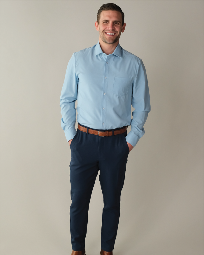 A man wearing a French blue shirt tucked into navy blue pants with his hands in the pants pockets with a plain beige background.