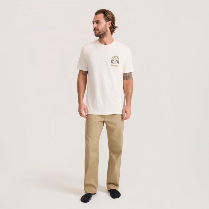 Man wearing cream colored Roaming Premium Tee with small logo on chest, paired with khaki pants and black socks