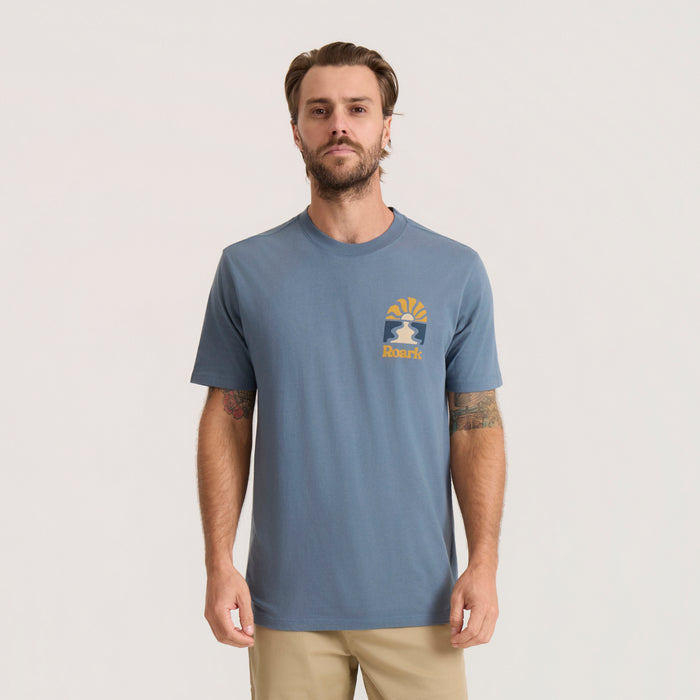 Slate blue short sleeve t-shirt with yellow and navy sunrise logo and Roark text on chest, worn by man against neutral background