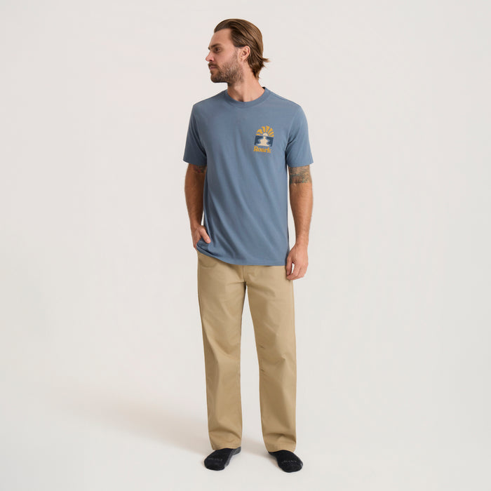 Man wearing slate blue Roaming Premium Tee with small chest logo, paired with tan pants, in full-body lifestyle shot