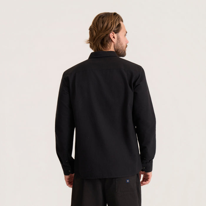 Man wearing black Scholar Chambray Button Up Shirt with long sleeves, shown from back view against neutral background