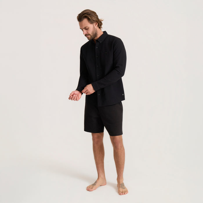 Man wearing black Scholar Chambray button-up shirt and black shorts, standing barefoot on neutral background