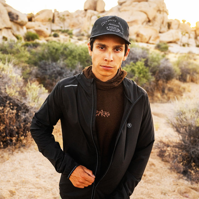Man wearing black Secondwind 3.0 jacket with brown turtleneck and cap in desert landscape setting