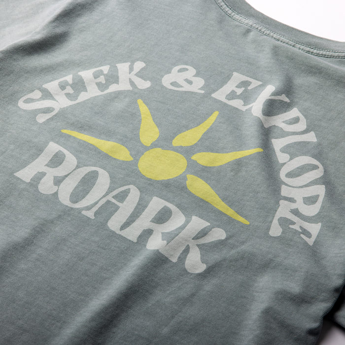 Hazy blue short sleeve tee with white "Seek & Explore" text and yellow sun graphic on chest, flat lay view
