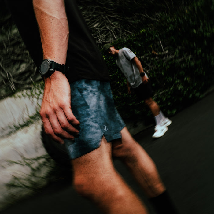 Man wearing navy tie-dye Serrano active stretch shorts during outdoor run on tree-lined path, detail view of shorts and watch