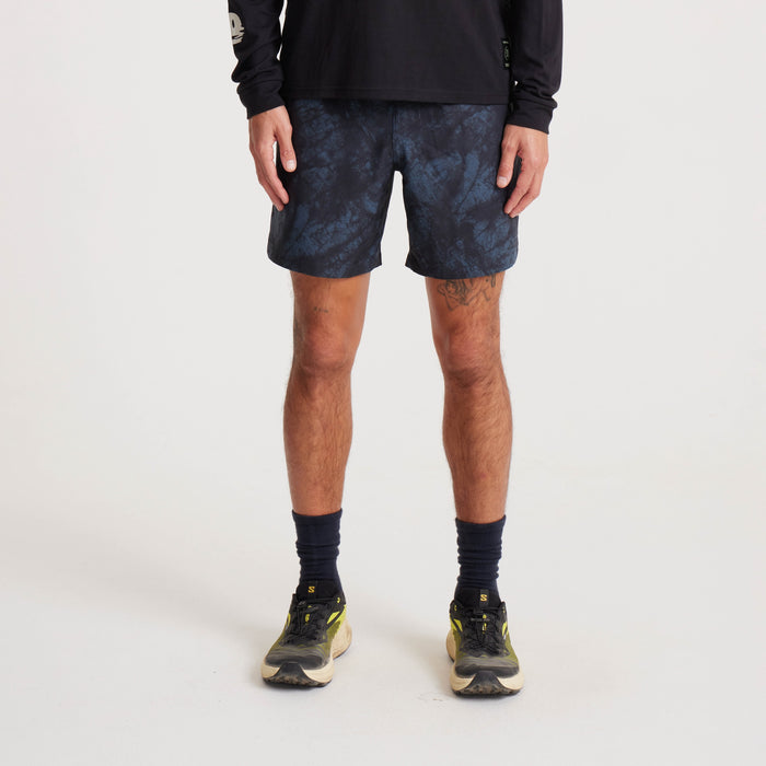 Model wearing navy tie-dye swim shorts with subtle blue patterns, paired with a black long-sleeve shirt and running shoes, showcasing a modern casual look.