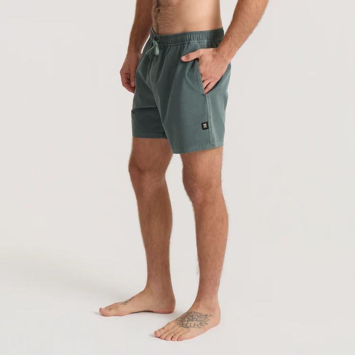 Man wearing sage green Shorey boardshorts with elastic waistband and side pockets, standing barefoot against neutral background