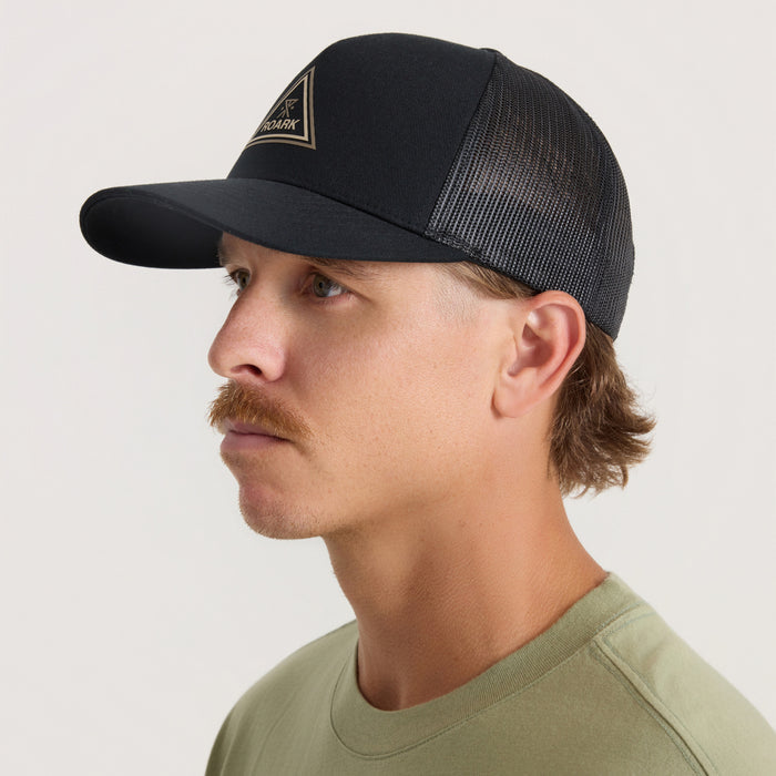 Black trucker hat with mesh back panel and gold triangle logo patch, worn by man in side profile view