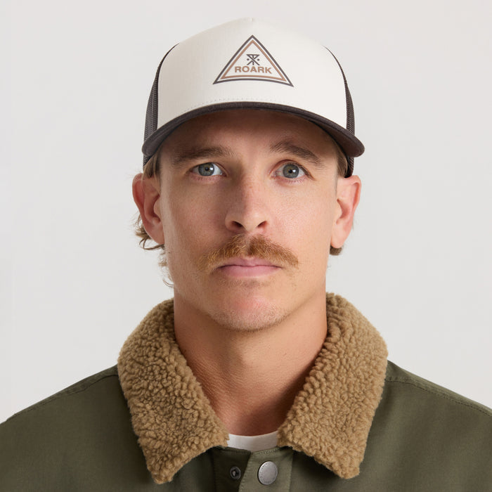 Man wearing cream and dark brown Roark Station Trucker Tricamp Snapback Hat with triangular logo patch on front