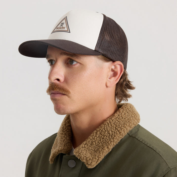 White and dark brown color-block trucker hat with mesh back and triangular Roark logo patch, worn by model in side profile view