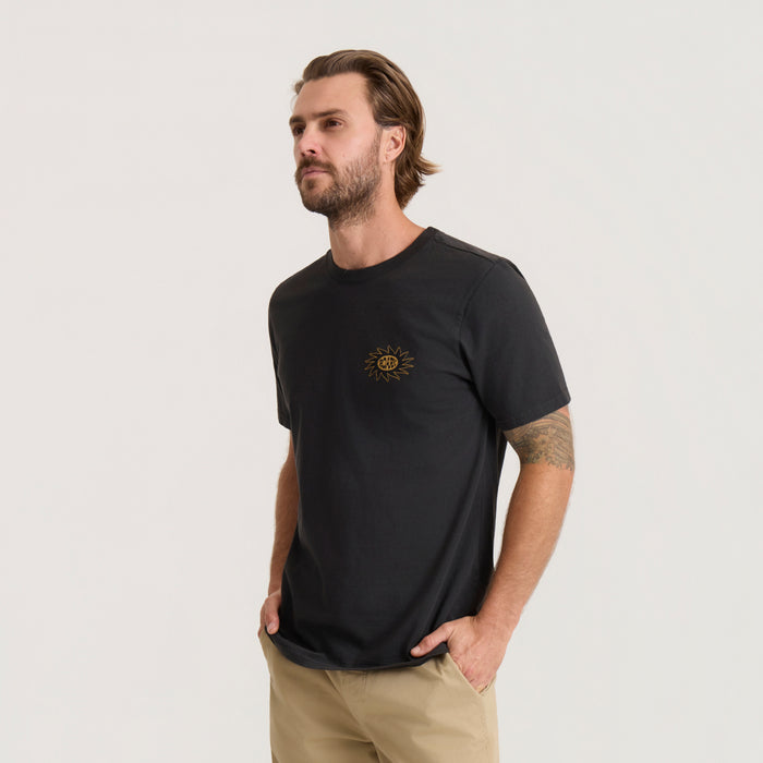 Black short sleeve t-shirt with small gold sun emblem on chest, worn by man standing against neutral background
