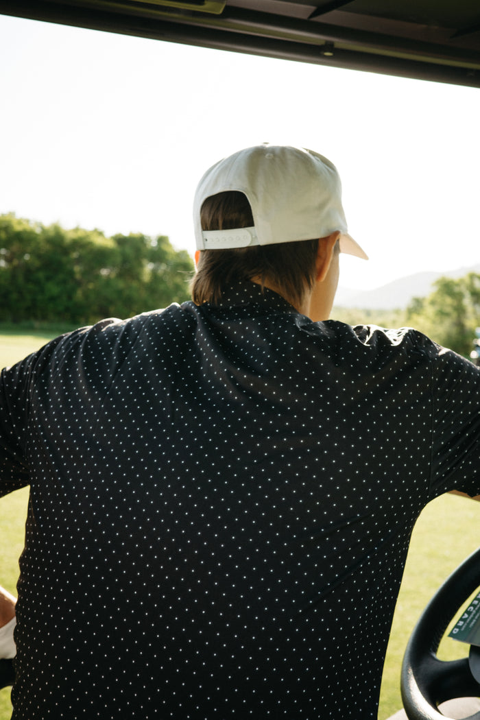 Person wearing a white cap and a black with white polka dots short sleeve polo shirt, looking out of a vehicle window with a scenic background.