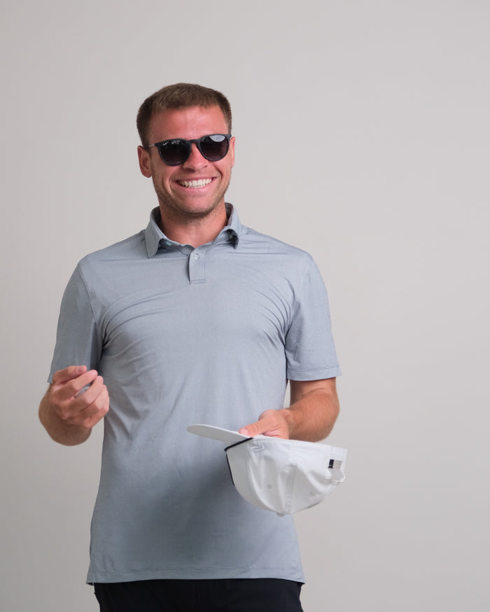 Man wearing a steel gray short sleeve polo shirt and sunglasses, holding a white cap against a gray background