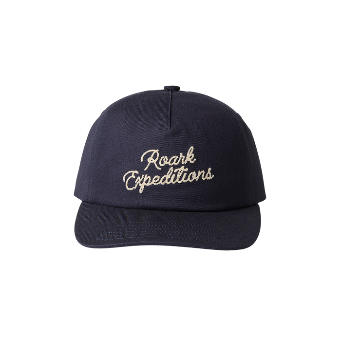 Navy snapback hat with cream embroidered Roark Expeditions script logo on front, flat lay view against white background