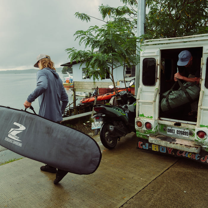 Person wearing Neptune blue Venture UPF hoodie with cap, carrying surfboard at waterfront dock with boats and tropical vegetation