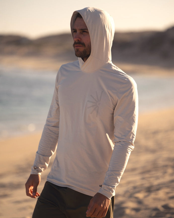 Bearded man wearing cream Venture UPF Hoodie with hood up, standing on beach at sunset