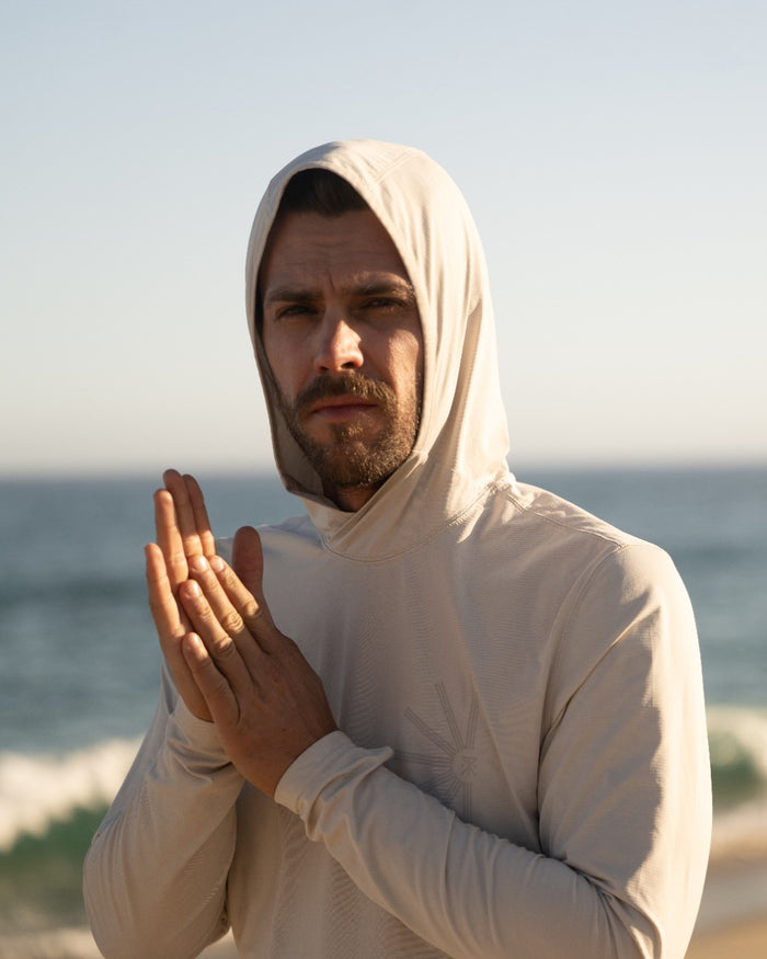 Bearded man wearing cream Venture UPF Hoodie with hood up, posed at beach with ocean background