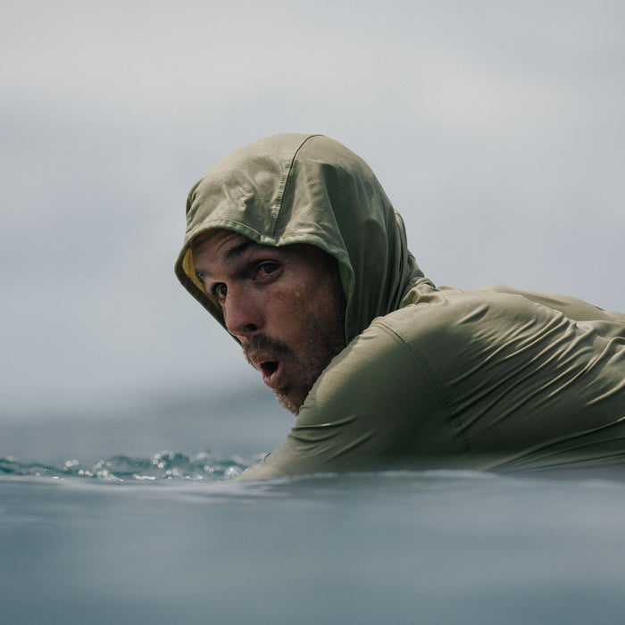 Man wearing sage green Venture UPF Hoodie with hood up while surfing in ocean water
