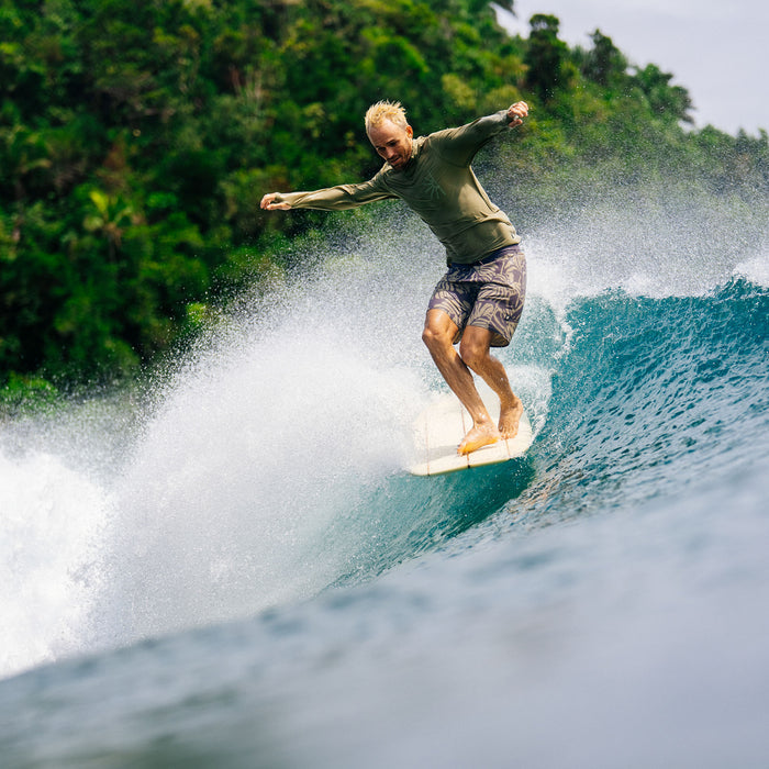 Man wearing sage green Venture UPF Hoodie surfing on white board in ocean wave with tropical trees background