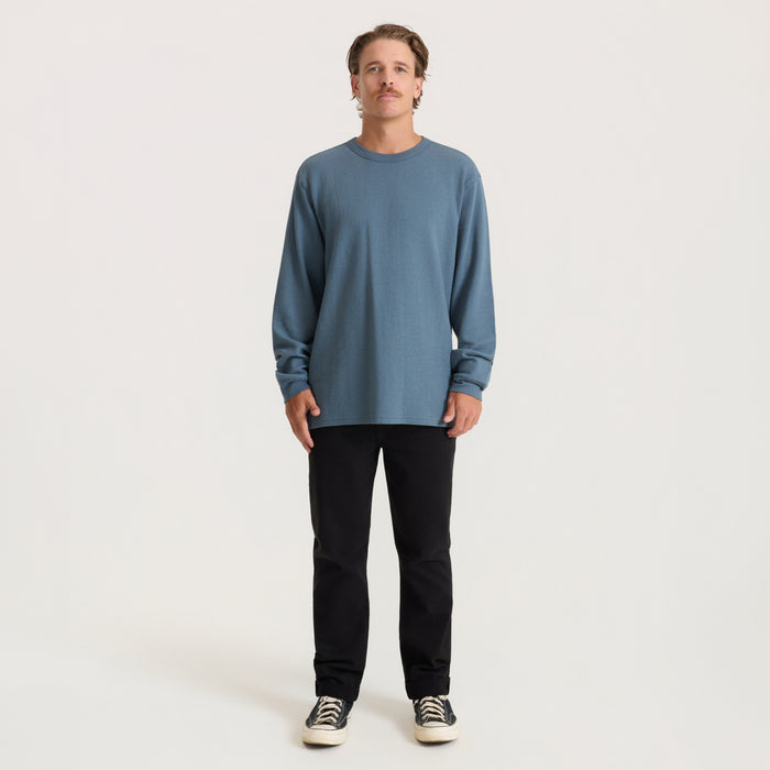 Man wearing slate blue well worn thermal long sleeve tee with black jeans and canvas sneakers, standing against neutral background