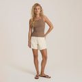 Woman wearing mocha brown sleeveless tank top with cream shorts and dark sandals, standing against neutral background