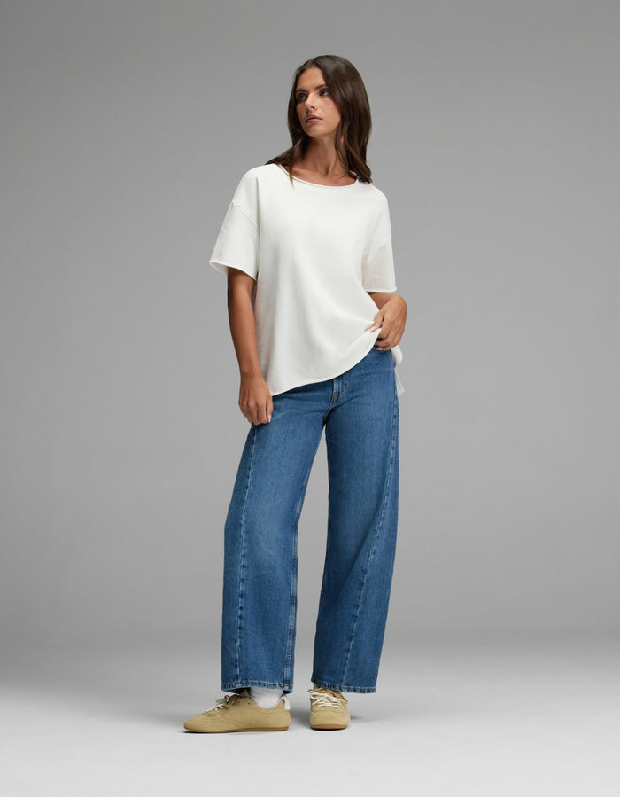 A woman stands against a gray background wearing the Cozy Earth Women’s Chelsea Short Sleeve, wide-leg blue jeans, and beige sneakers, with one hand in her pocket and looking to the side.|Color:Bone