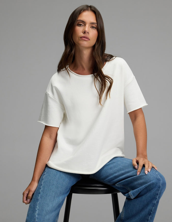 A woman with long brown hair, wearing the Cozy Earth Women’s Chelsea Short Sleeve and blue jeans, sits on a black stool against a plain gray background, looking confidently at the camera.|Color:Bone