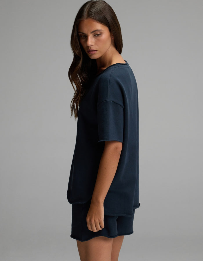 A woman with long brown hair stands sideways against a plain gray background, wearing the Cozy Earth Women’s Chelsea Short Sleeve top and matching shorts in dark blue. |Color:Eclipse