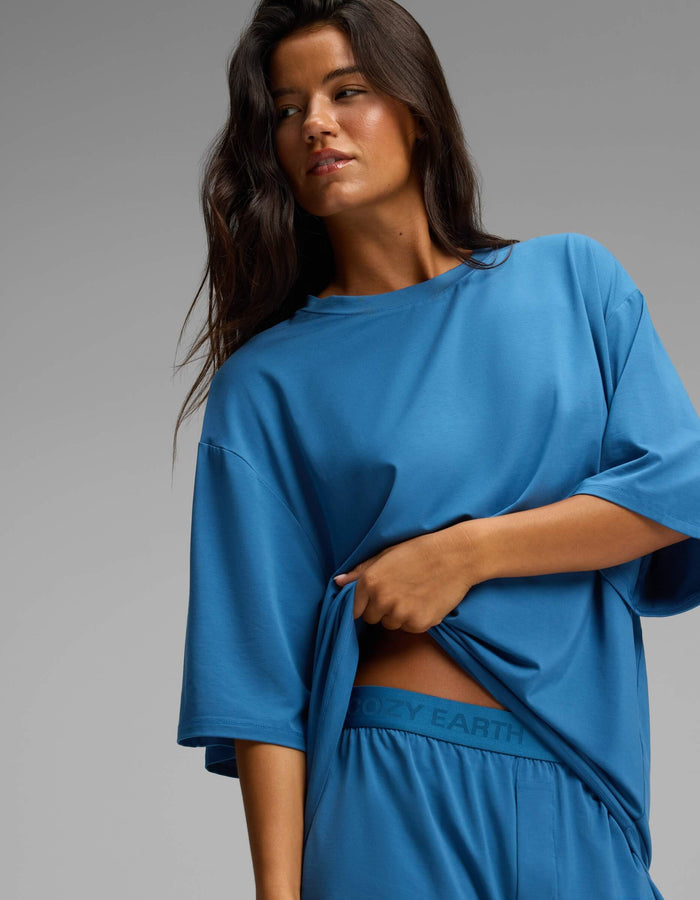 A woman with long dark hair gazes to the side against a plain gray background while lifting the hem of her loose blue Cozy Earth Women’s First Layer Boyfriend Sleep Tee to reveal matching blue Boxer Shorts featuring the “COZY EARTH” logo on the waistband.|Color:Anchor