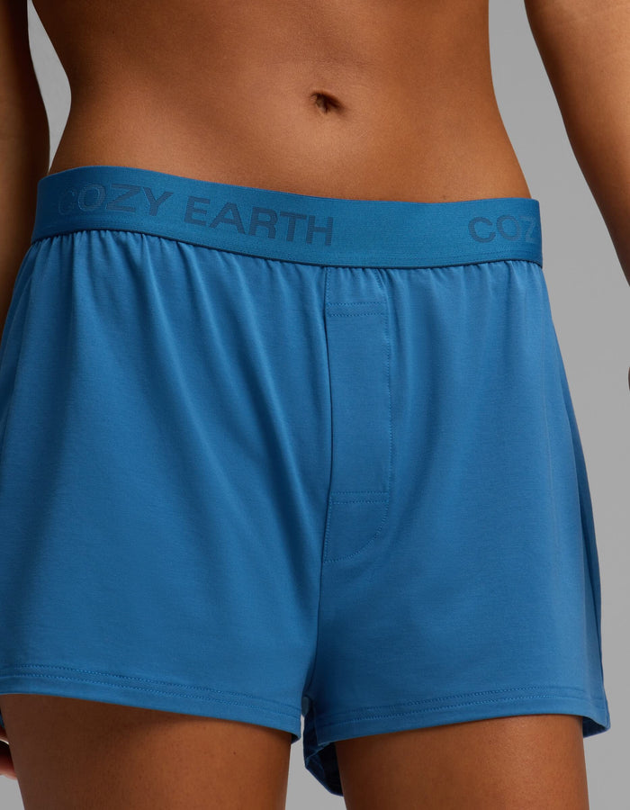 A person models the Cozy Earth Women’s First Layer Boyfriend Sleep Tee & Boxer Short Set, featuring blue boxer shorts with the brand name “Cozy Earth” printed on the waistband, against a neutral background. Only the torso and upper legs are visible.|Color:Anchor