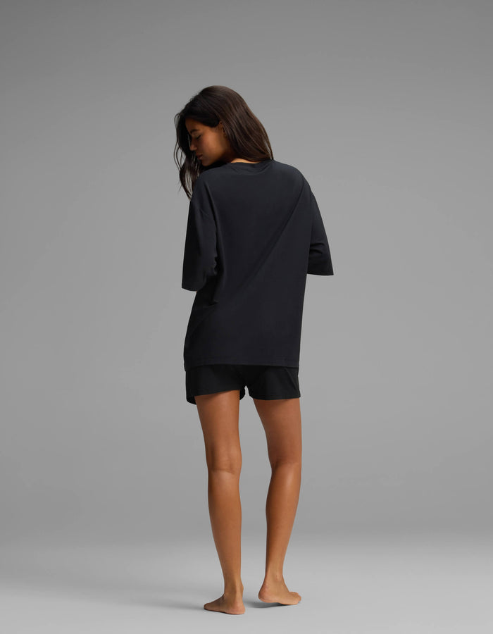 A person stands barefoot on a gray floor, facing away from the camera, wearing the Cozy Earth Women’s First Layer Boyfriend Sleep Tee & Boxer Short Set in black against a plain gray background.|Color:Jet Black